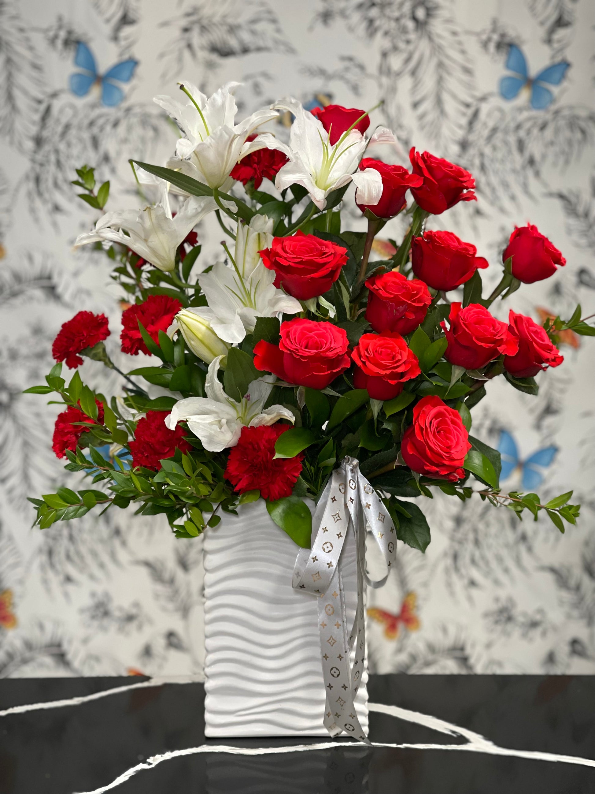 Elevate your floral experience with the Audrey Vase, a captivating arrangement that embodies elegance and luxury. This stunning display features one dozen Nina Scarlet Roses, complemented by imported White Rose Lilies and vibrant Red Carnations, all beautifully intertwined with delicate, fresh foliage. Each element is carefully chosen for its quality and visual impact, creating a harmonious and eye-catching bouquet.