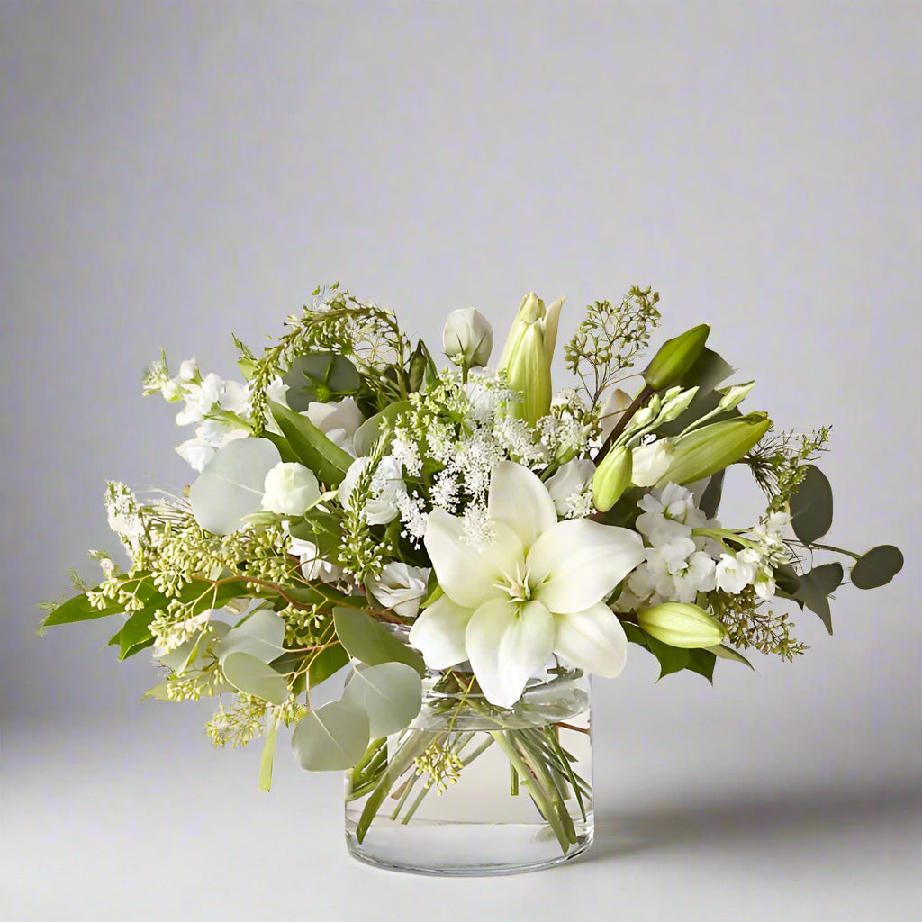 The Silk Sympathy Vase is a timeless and elegant sympathy flower arrangement, designed to offer comfort and support during times of loss. This small yet meaningful floral display features a delicate blend of white lilies, spray roses, stock, eucalyptus, and Queen Anne’s lace, arranged in a classic clear glass 5x5 vase. Perfect as a condolence gift, this arrangement brings a sense of peace, purity, and remembrance to any setting.