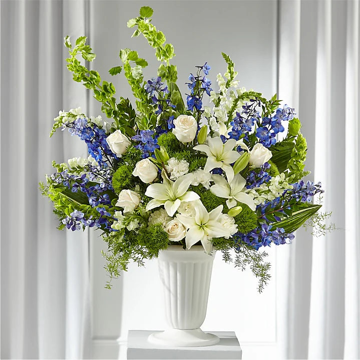 Express your condolences with the Sapphire Sympathy Pedestal Arrangement, a stunning floral tribute designed for funeral services, memorials, and sympathy gifts. This large, elegant arrangement is displayed in a classic pedestal vase, featuring a harmonious blend of white, blue, and green blooms, symbolizing peace, remembrance, and serenity.

Features:
Premium white lilies, snapdragons, and roses for purity and grace
Blue delphinium to add depth and tranquility